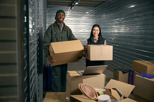 Two people holding moving boxes inside a self storage unit aisle with stacked boxes and a laptop nearby. 